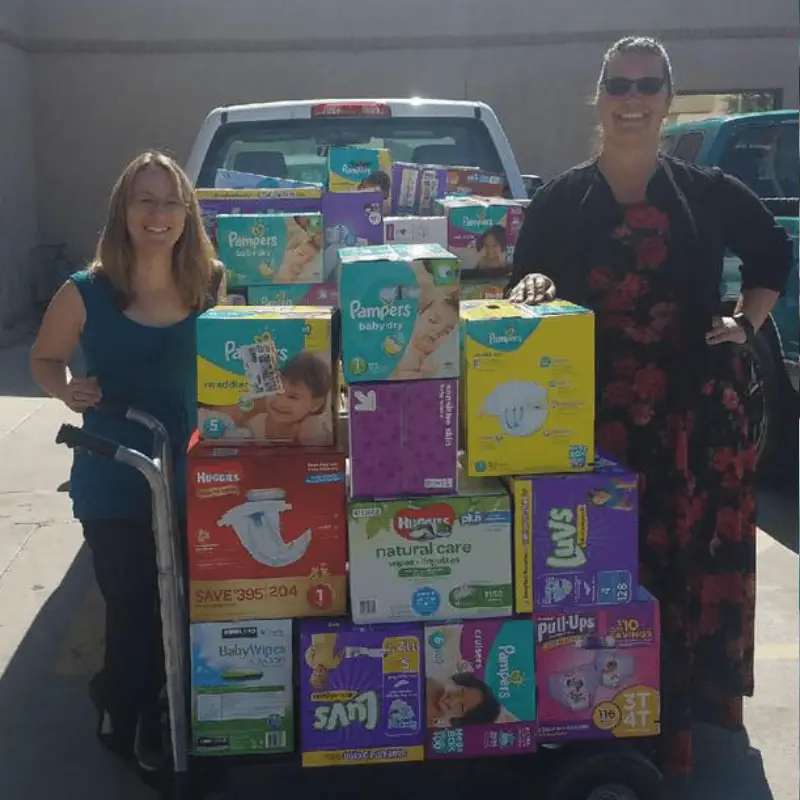 Two women stand beside a cart loaded with various brands of diapers and baby supplies, in front of a pickup truck.