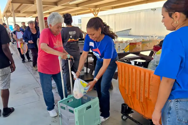 A volunteer hands a bag of food to an elderly woman using a walker, while others wait in line at a community food distribution event.